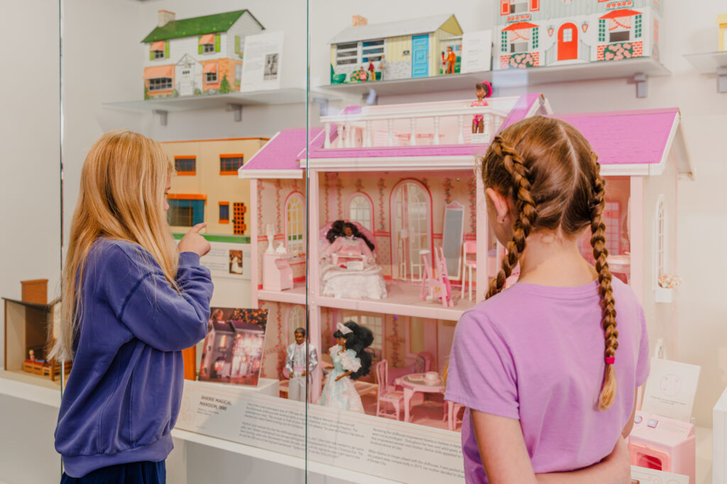 two young girls consider a dollhouse on display behind a glass case