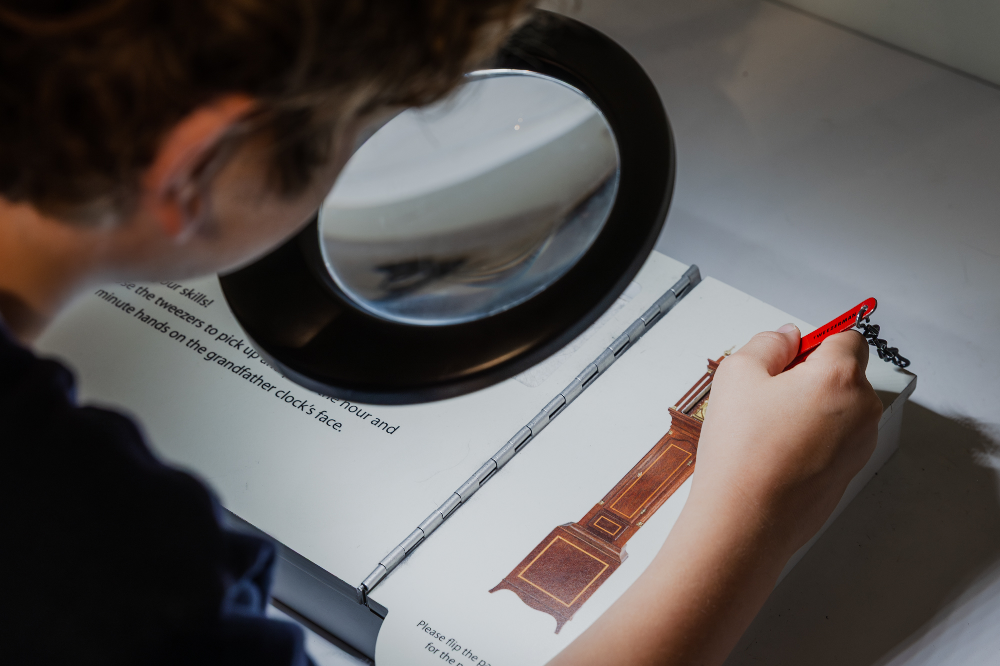 a child looks through a magnifying glass, using tweezers to move the hands on a miniature clock