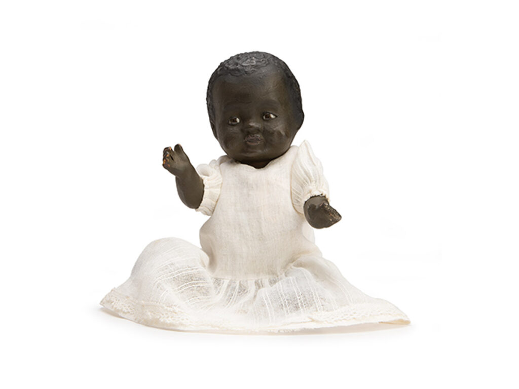 Black baby doll with a paper mache head wearing a white dress.