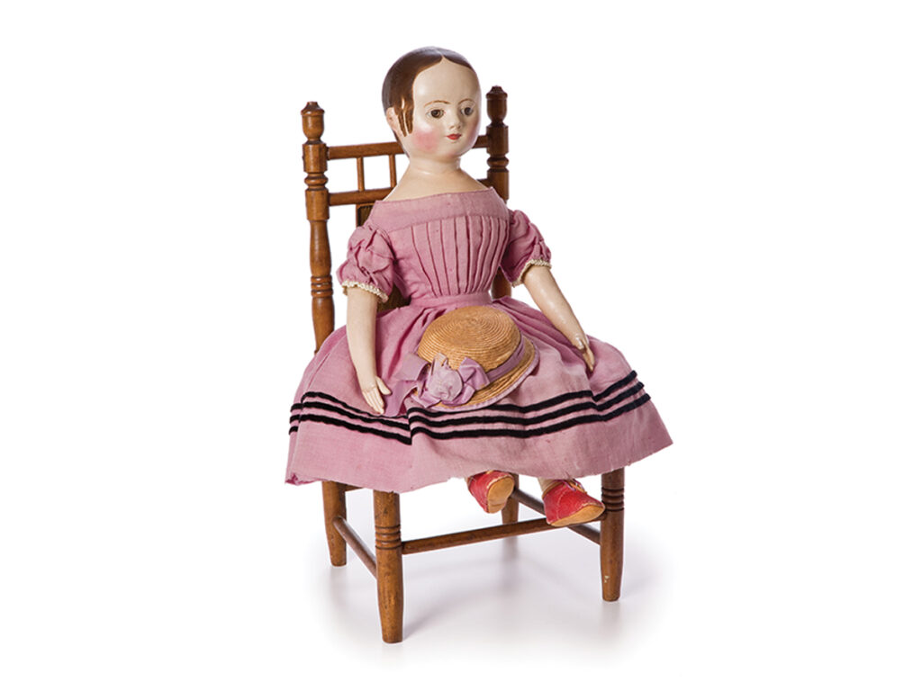 Doll in a pink dress holding a hat and sitting on a wooden chair.