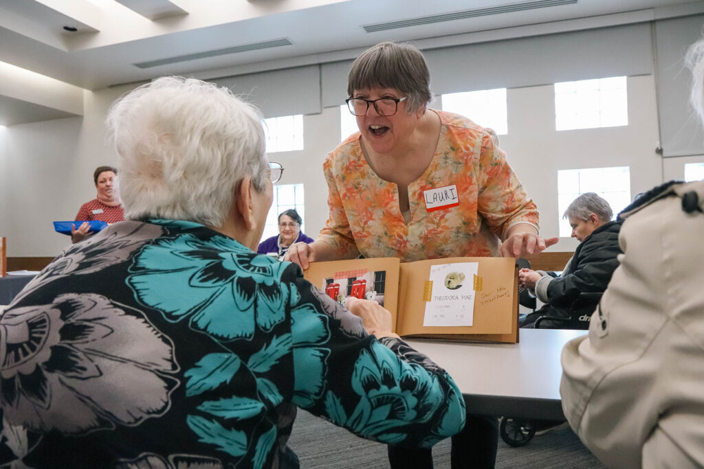 An older woman is shown a book of miniature objects.